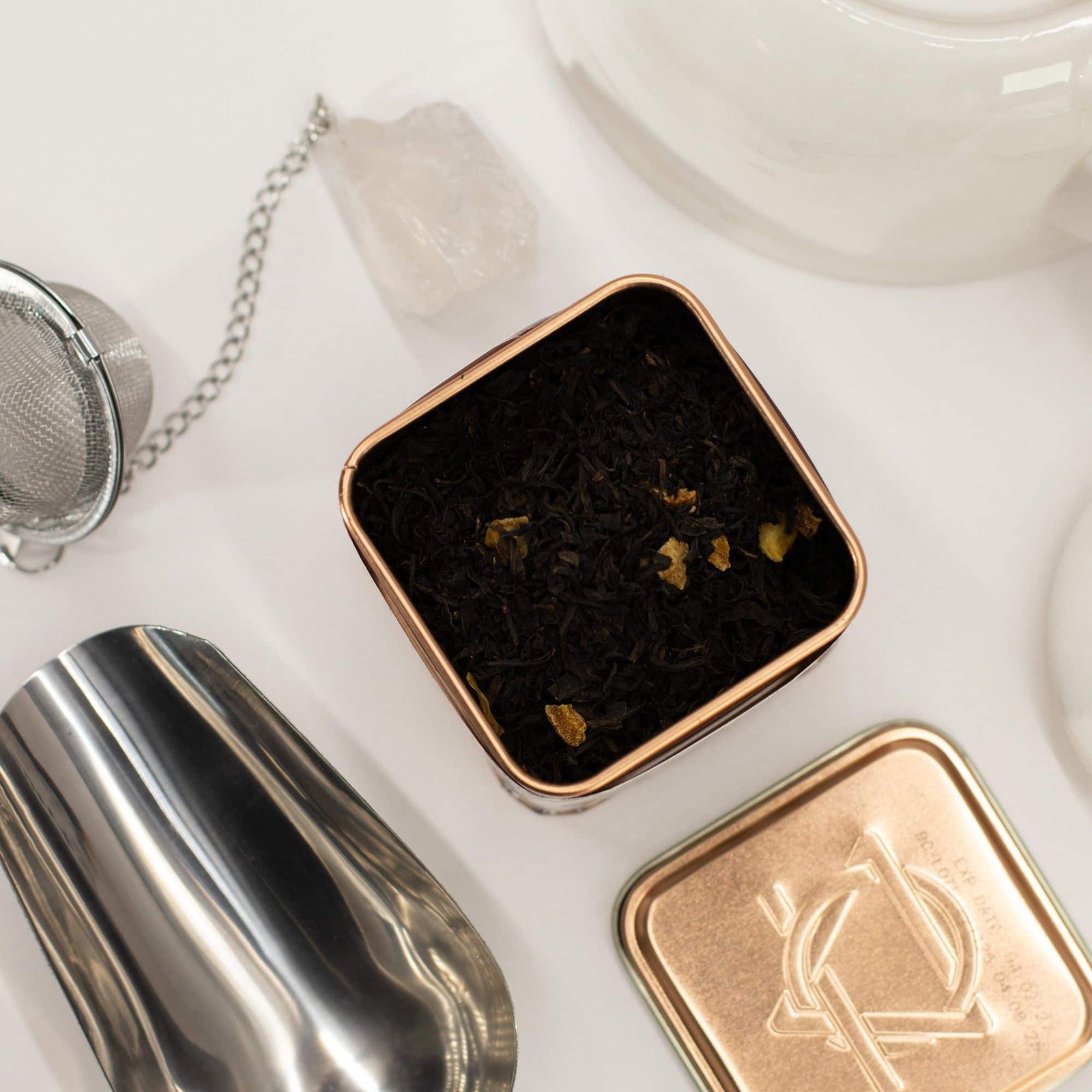Close-up of loose black tea leaves in a decorative tin with gold accents, surrounded by white ceramics and a crystal, on a white background.