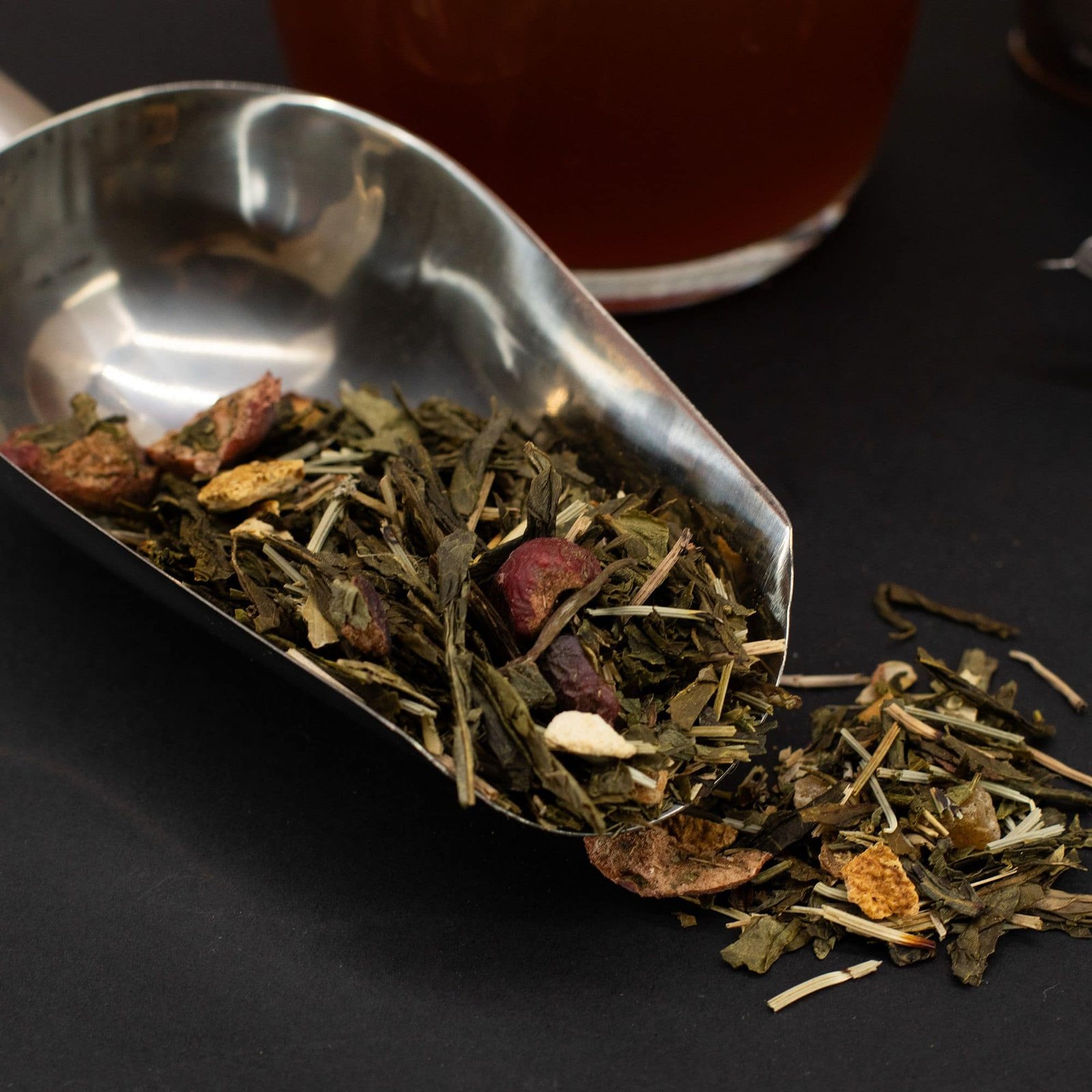 Close-up of a herbal tea blend with dried herbs and flowers in a metal scoop, on a black surface.