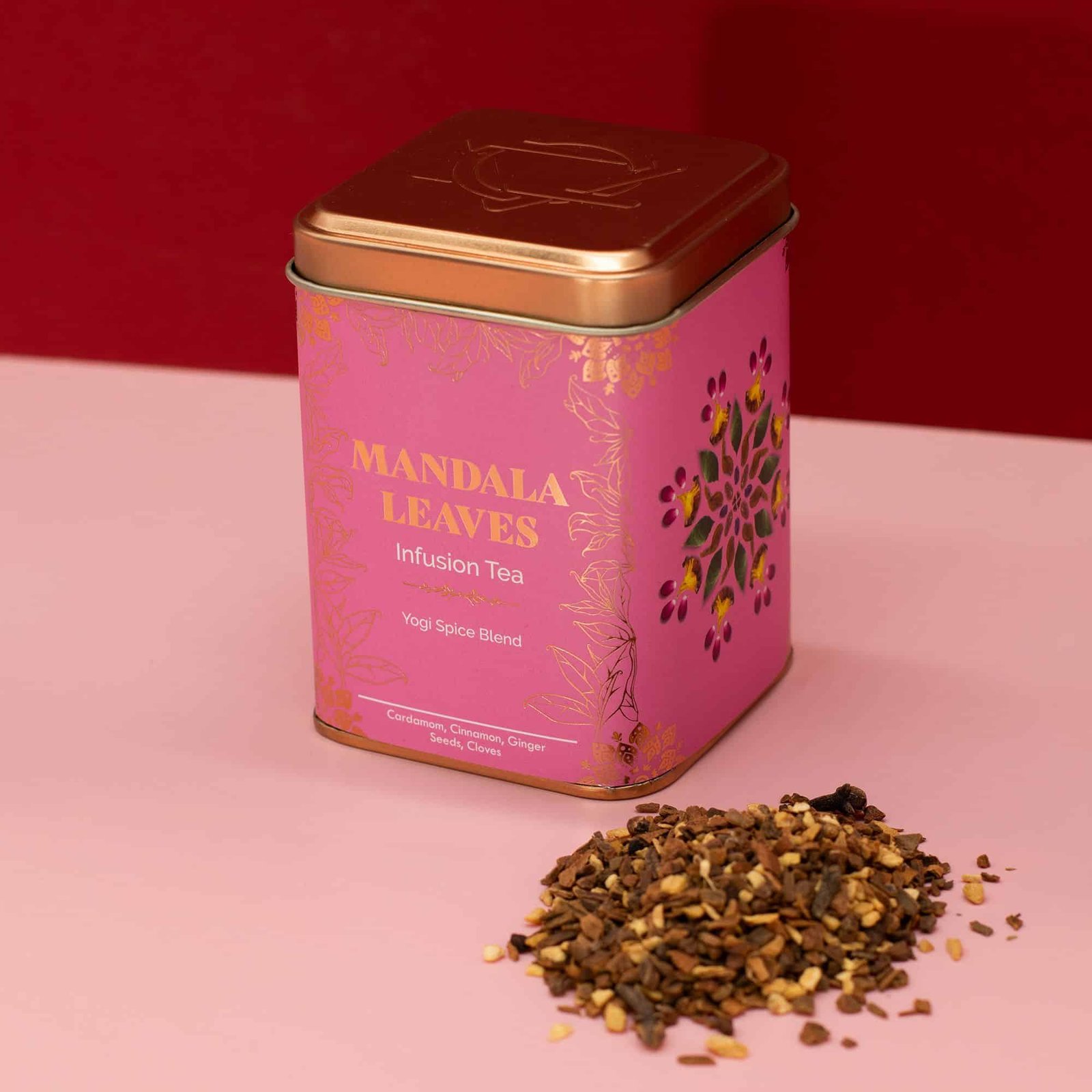 A pink metal tin of Mandala Leaves infusion tea with a floral design, accompanied by loose tea leaves on a pink surface.