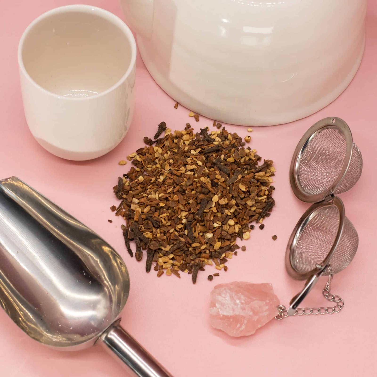 Close-up of herbal tea ingredients with a tea strainer, cup, and crystal on a pink background, showcasing natural wellness products.