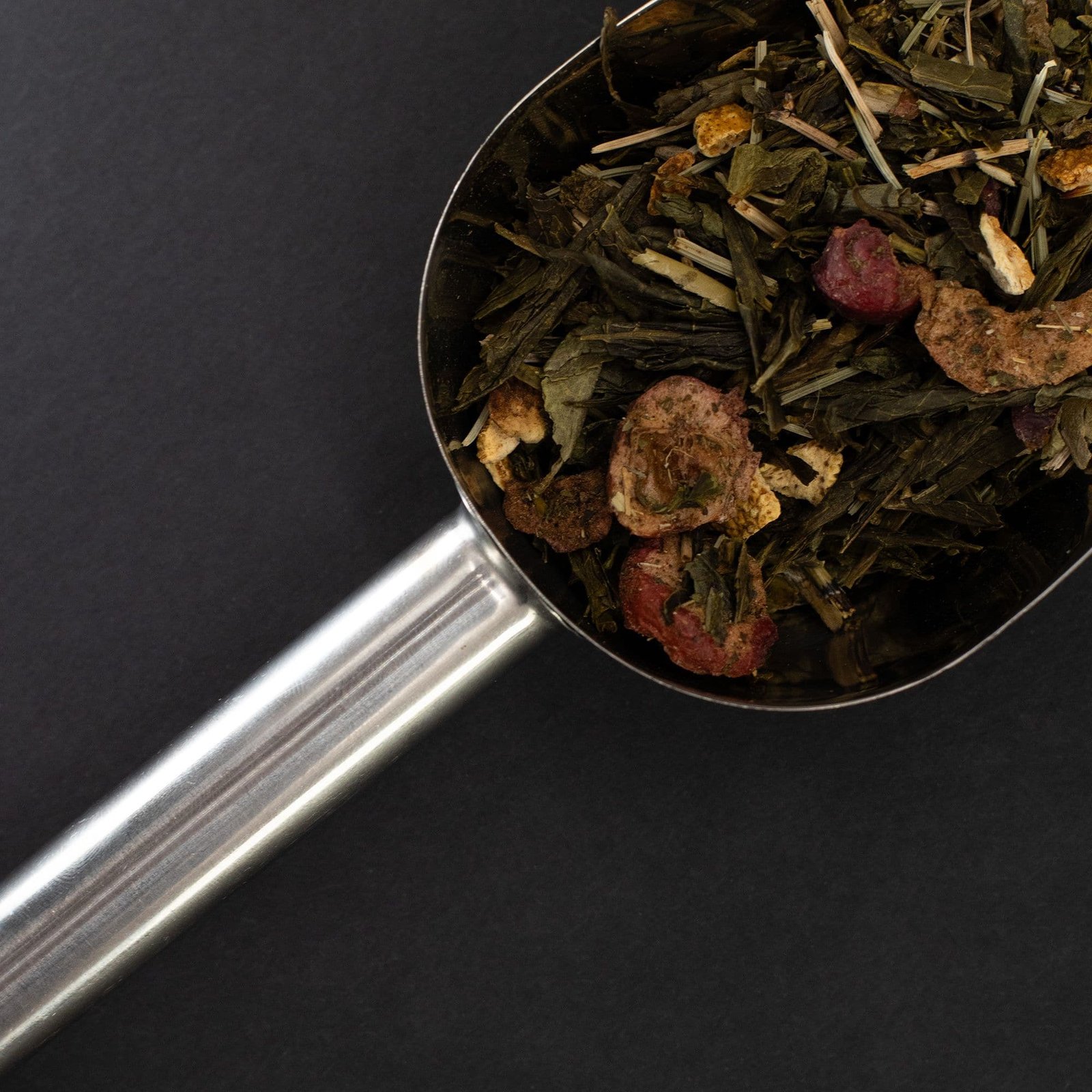 Close-up of a stainless steel scoop filled with loose herbal tea leaves, dried herbs, and berries on a dark background. Perfect for herbal tea enthusiasts and gift buyers.
