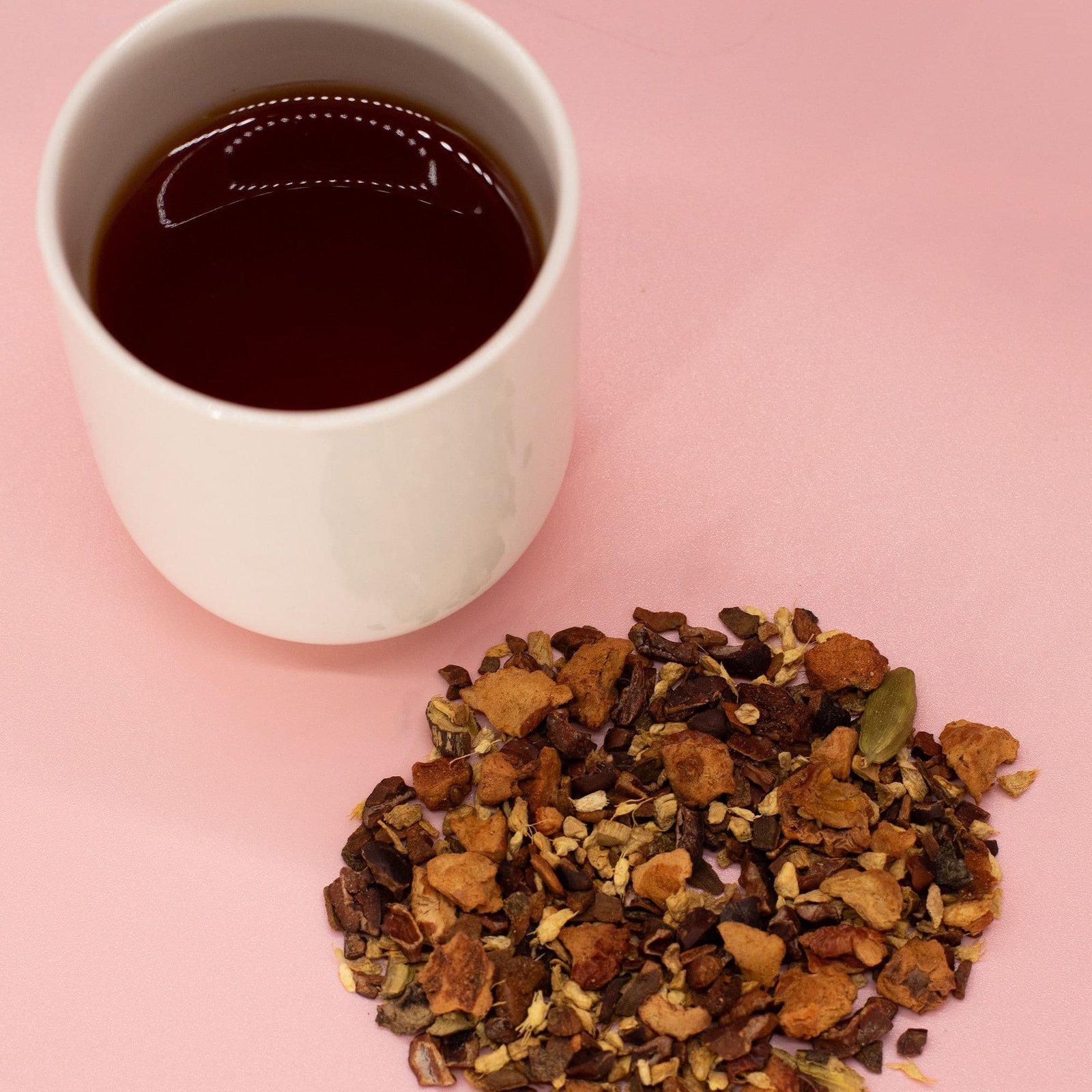 White ceramic mug filled with hot tea or coffee on a pink background, with loose herbal tea leaves scattered nearby, perfect for gift ideas.