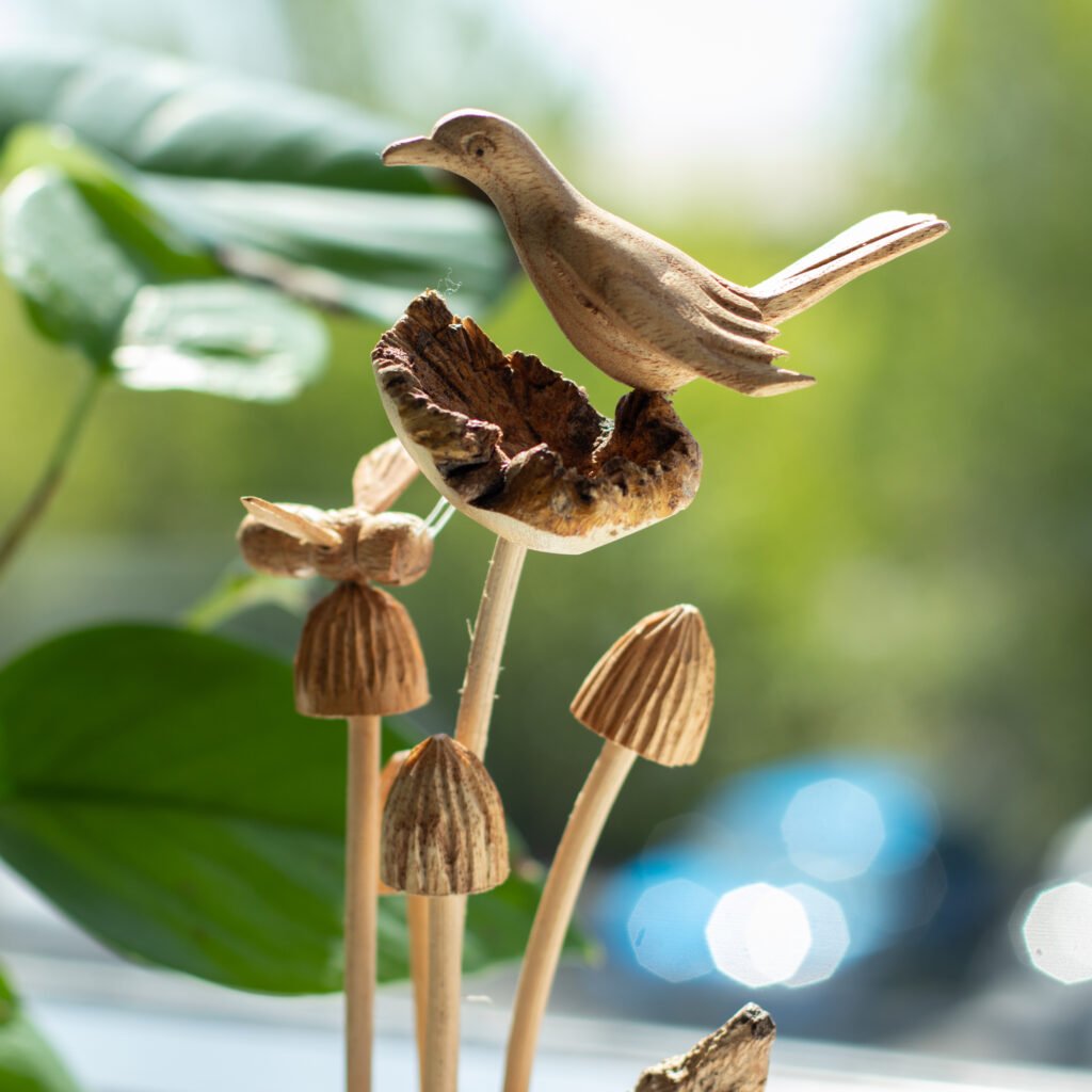 Handcrafted parasite wood bird sculpture on natural wood mushrooms with blurred greenery in the background.